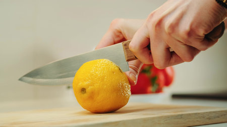 Close-up shot of hands with knife cutting lemon on the kitchen board.
