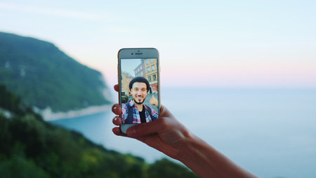 Hand holding smartphone during video call with man in front of the sea. Beautiful landscape in the...