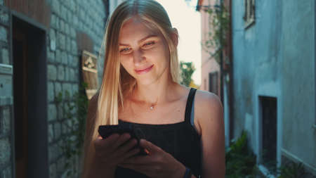 Close-up of positive blonde woman reading something on smartphone while walking the street. She happy and smiled. Summer time.の写真素材