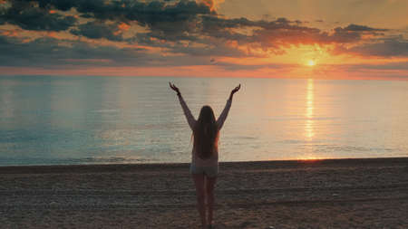 Woman with long hair walking to the sea and raising her hands up. Looking on sunset. Back view.の写真素材