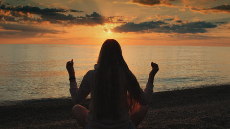 Back view of woman practicing yoga in meditation pose on the beach. Beautiful sunset on the sea.の写真素材