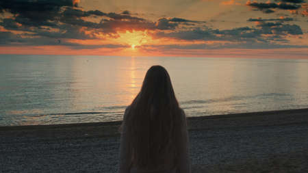 Close-up shot of woman with long hair walking to the sea and raising her hands up. Looking on sunset. Back view.の写真素材