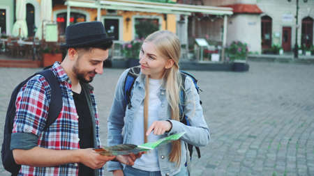 Trendy couple of tourists finding necessary destination on map and admiring surroundings. They standing on city square.の写真素材