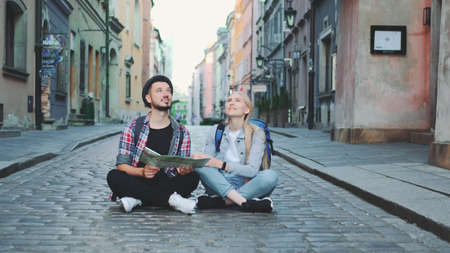 Young tourists couple using map, sitting on pavement and admiring historical surroundings. They are excited and smiled.の写真素材