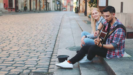 Young couple of street singers in old part of the city sitting on sidewalk. Man playing guitar and woman singing.の写真素材