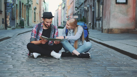 Young tourists couple using map, sitting on pavement and admiring historical surroundings. They are excited and smiled.の写真素材