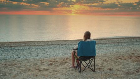 Woman sitting on camping chair and drinking coffee. Relaxing and enjoying nature. Admiring sunrise on the sea.の写真素材