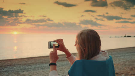 Young woman making video of sunset with smartphone sitting on the seashore. Back viewの写真素材