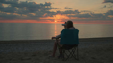 Back view of woman sitting on camping chair and drinking coffee. Relaxing and enjoying nature. Admiring sunrise on the sea.の写真素材