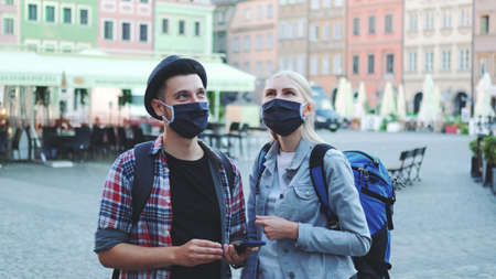 Man and woman tourists in protective masks and with bags using smartphone. They looking for interesting tourist attractions.の写真素材
