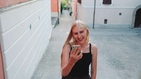 Pretty blonde woman sharing her impressions from summer trip on video call on smartphone while walking the city of her visitの写真素材