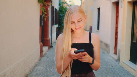 Pretty blonde girl chatting on smartphone while walking down the street of the old town. Smiling and happy woman.の写真素材
