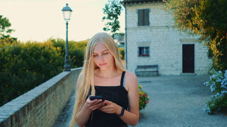 Charming young woman using smartphone while walking outside in summer. She reading something and chatting.の写真素材