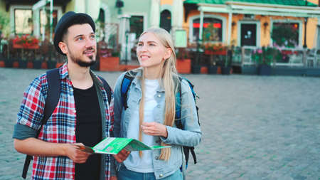 Trendy couple of tourists finding necessary destination on map and admiring surroundings. They standing on city square.の写真素材