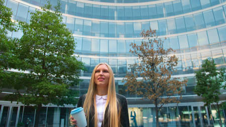From below of serious blond executive woman in suit having coffee in paper cup looking away on street.の写真素材