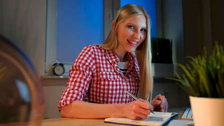 Lively young blond woman in casual checkered shirt, sitting at wooden desk with notebook holding metal pen and looking at camera smiling at night in dark room.の写真素材