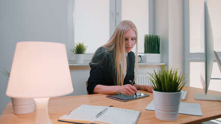 Attractive blond female with long hair in elegant suit, sitting at light wooden desk with computer and concentrating on work on tablet with pen in light office with plants.の写真素材