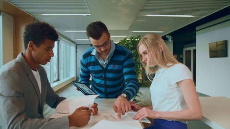 Side view of black man with coworkers at table reading papers and using tablet in team at table.の写真素材