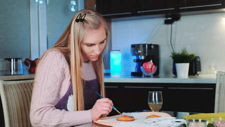 Pretty young woman learning to decorate gingerbread cookies at home. She sitting at the table and painting.の写真素材