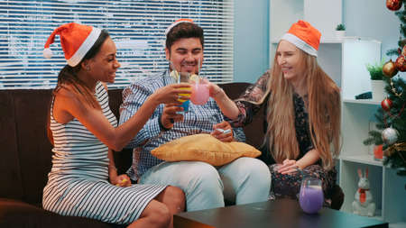 Beautiful young people in Santa hats making cheers and blowing party whistle. They sitting on sofa, smiling and drinking. Friends happiness conceptの写真素材