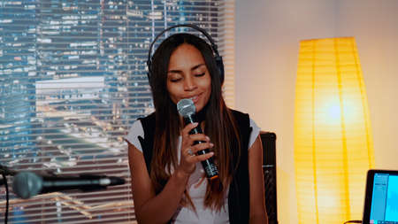 Close-up of beautiful multiracial girl in headphone singing into microphone and recording a song in home studio with skyscrapers in the background.の写真素材