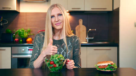 Young lady preferring salad to hamburger. Attractive young woman choosing to eat healthy salad for lunch while sitting at table in stylish kitchen and doing thumbs up gesture and looking at camera.の写真素材