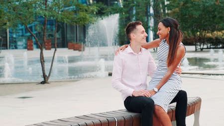 Romantic multiracial couple having a date: woman whispering something in boyfriends ear. They sitting on a bench, embracing and kissing.の写真素材