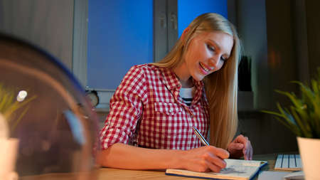 Lively young blond woman in casual checkered shirt, sitting at wooden desk with notebook holding metal pen and looking at camera smiling at night in dark room.の写真素材