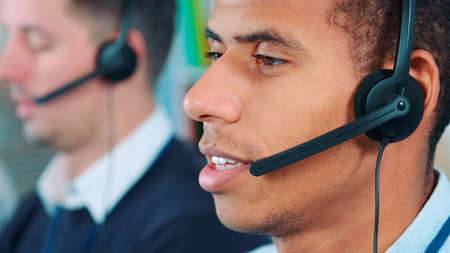 Close-up shot of mixed-race man in headset talking to somebody in office. In the background there is his colleague working.の写真素材