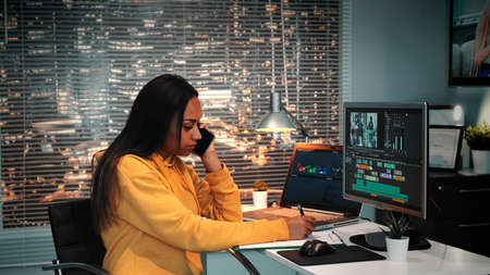 Mixed race colorist talking by smartphone and writing notes in modern production studio. In the backgrounds - skyscrapers.の写真素材