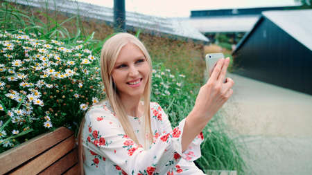 Woman making video call on green roof. Lovely young female using smartphone to make video call while sitting on bench on living roof of huge mall in USA.の写真素材