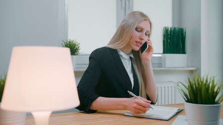 Elegant young blond female in office suit sitting at workplace with computer and negotiating via mobile phone in hand writing down necessary information into notebookの写真素材
