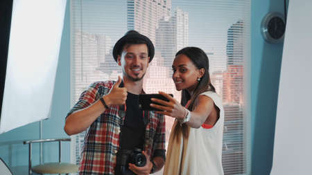 Beautiful african model making selfie with handsome photographer in professional studio photo shoot. There is soft box in the background.の写真素材