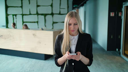 Stylish blond woman in jacket standing in contemporary office hall and surfing smartphone with smileの写真素材