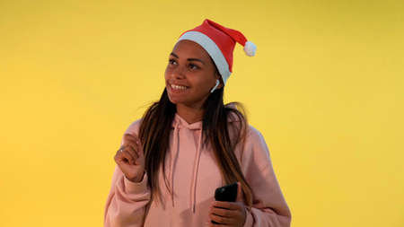 Happy african woman in Santa hat and wireless headphones listening to the music on yellow background. She singing some Christmas songs.の写真素材