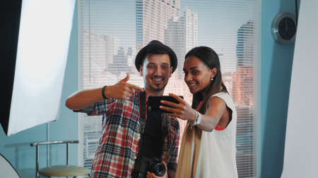 Beautiful african model making selfie with handsome photographer in professional studio photo shoot. There is soft box in the background.の写真素材