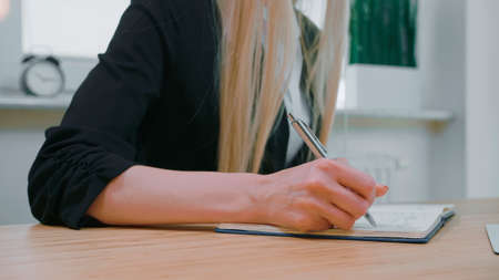 Crop view of woman with long blond hair in bright casual shirt, sitting at wooden desk and writing down information with shiny metal pen into daily planner at day.の写真素材