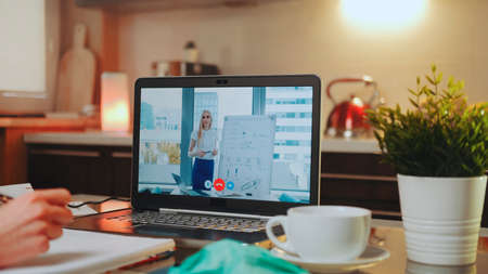 Online video conference on laptop with female speaker in home office. In the foreground: writing hands, medical mask and a cup.の写真素材