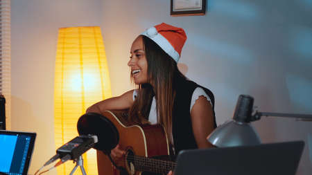 Beautiful black woman in Santa hat singing and playing guitar on Christmas Eve in cozy home studio. In front there is a recording equipment.の写真素材