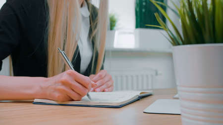 Crop view of woman with long blond hair in bright casual shirt, sitting at wooden desk and writing down information with shiny metal pen into daily planner at day.の写真素材