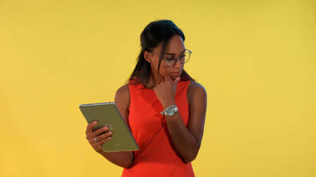 Black business woman in dress and eyeglasses analysing something on tablet There is yellow background.の写真素材