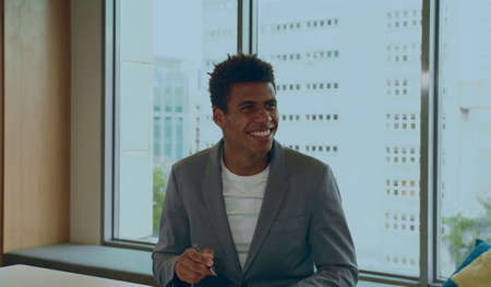 Cheerful elegant African-American man in suit with pen smiling while communicating with coworkers in officeの写真素材