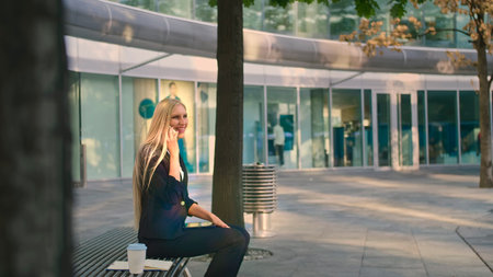 Elegant modern woman sitting on bench and waving with hand to friend while speaking on smartphone outdoorsの写真素材