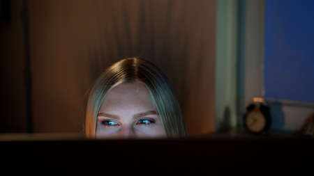 Crop view of beautiful female sitting at night at computer and looking at camera over top of monitor with dark window on blurred backgroundの写真素材