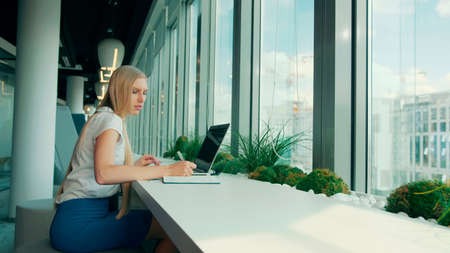 Side view of businesswoman writing on paper while sitting with laptop at table near window in modern officeの写真素材