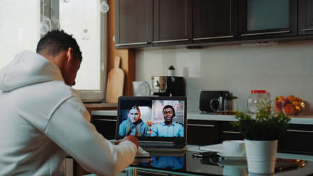 Multiracial colleagues working home on quarantine by making common video call by computer. Black man sitting in the kitchen and writing.の写真素材