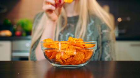 Close-up shot of woman eating potato chips. Beautiful young female enjoying potato chips sitting in stylish kitchen.の写真素材