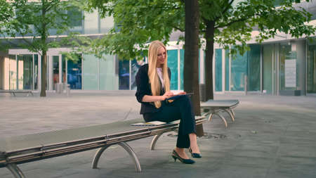Formal business woman sitting on bench in office patio and browsing tablet in leisure.の写真素材