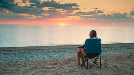 Woman admiring sunset on the sea sitting in folding tourist chair. Relaxing and enjoying nature.の写真素材