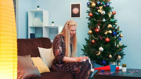Pretty woman writing a postcard near the Christmas tree. She is sitting on sofa near table and big standing lamp. Side view.の写真素材